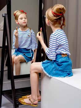 A nice little girl near the big mirror paints her lips with lipstick. The concept of childhood, style and fashion.