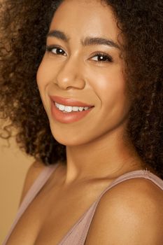 Face closeup of cheerful mixed race young woman with curly hair smiling at camera while posing, standing isolated over light background. Natural beauty, skincare concept