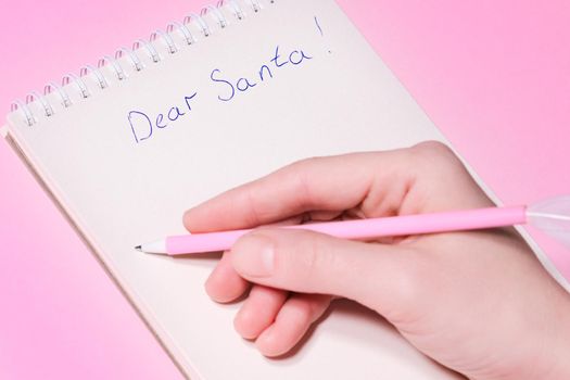 Dear Santa letter concept. Hand holding pink pen decorated with feather and writing in Notepad letter to Santa Claus on pink background