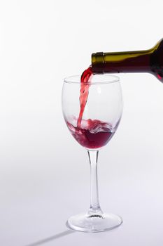 Red wine pouring from bottle into big glass on white background.