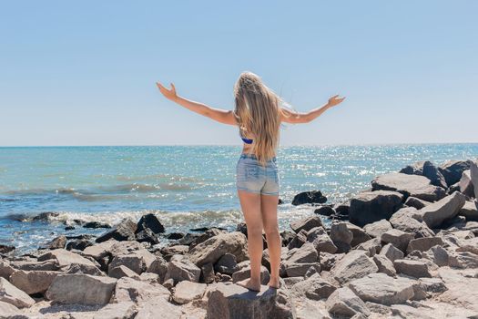 A young teenage girl with long blonde hair blonde in short shorts raised and spread her hands to the sides standing on the rocks near the sea shore and the beach. The concept of freedom.