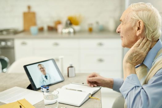 Side-view photo of distressed elderly man touching his neck while having an online video consultation with medical specialsit