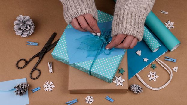 Close up woman hands packing and wrapping Christmas gift boxes with blue paper, adding envelope and skeleton leaves, high angle view