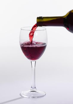 Red wine pouring from bottle into big glass on white background.