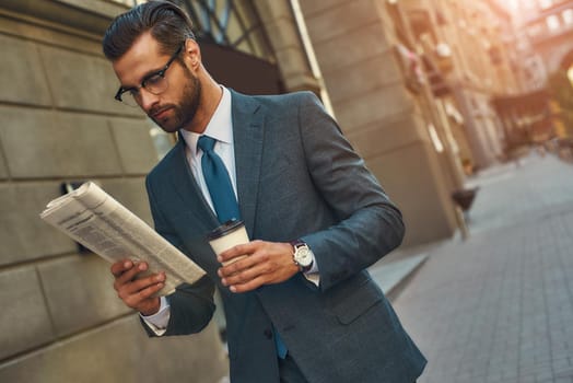 Young handsome bearded businessman in eyeglasses reading newspaper and holding cup of coffee while walking outdoors. Media. Information