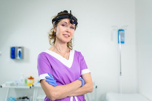 Medical worker of ENT clinic middle aged Caucasian female in purple medical uniform posing looking at the camera in the examination room. Professional medical specialist otolaryngologist in hospital.