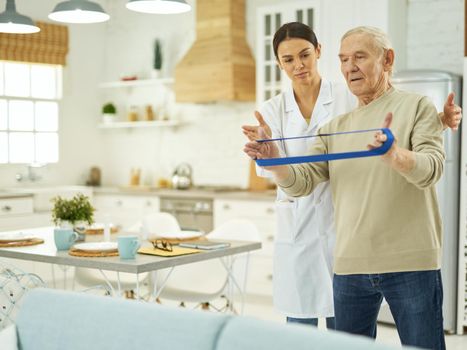 Young female doctor in a white coat explaining to a pensioner how to do exercises at home with fitness rubber