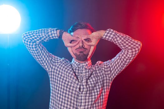 Funny positive young hipster guy in a plaid shirt pranks while depicting his glasses on a blue-red background. Concept of a cheerful young man