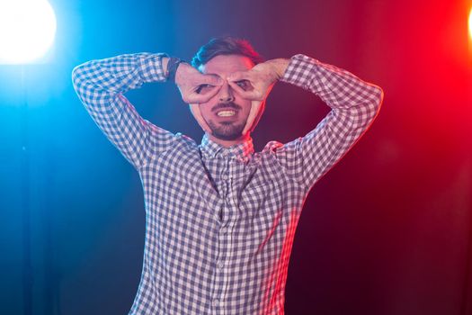 Funny positive young hipster guy in a plaid shirt pranks while depicting his glasses on a blue-red background. Concept of a cheerful young man