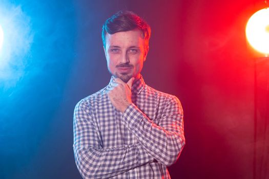 Carming young smiling hipster man is posing against the background of red and blue studio lighting. Concept of pensive man
