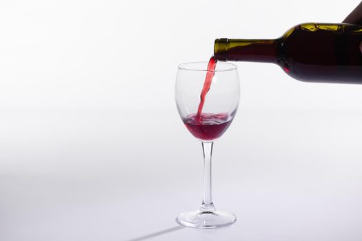 Red wine pouring from bottle into big glass on white background.