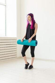 Slim young mixed race girl hipster with colored hair folds gymnastic mat after yoga in the gym. Healthy lifestyle and fitness concept