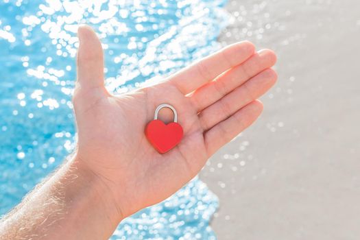In the hand of a man on the palm of a small red heart close-up in the form of a lock. A sign or symbol of love against the sea coast.