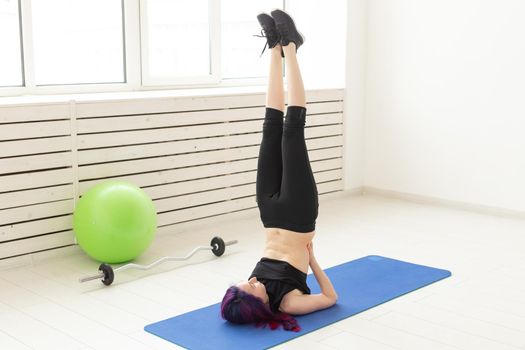 Young slim girl with colored hair makes an headstand on a sports blue mattress next to a barbell and green fitball. Concept of good physical fitness and regular training in the gym