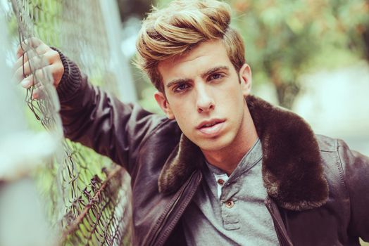 Portrait of a young handsome man, model of fashion, with modern hairstyle in urban background wearing aviator leather jacket. Blonde hair