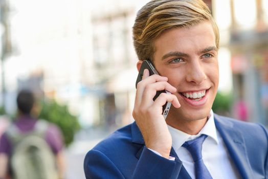 Portrait of an attractive young businessman on the phone in urban background, wearing blue suit and tie. Blonde hair