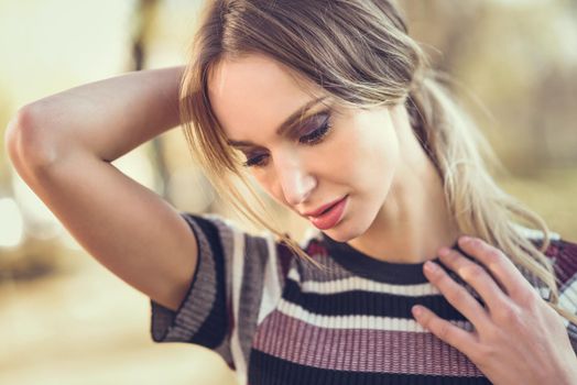Close-up portrait of pensive young blonde woman standing in the street. Beautiful girl in urban background wearing striped sweater. Female with straight hair.