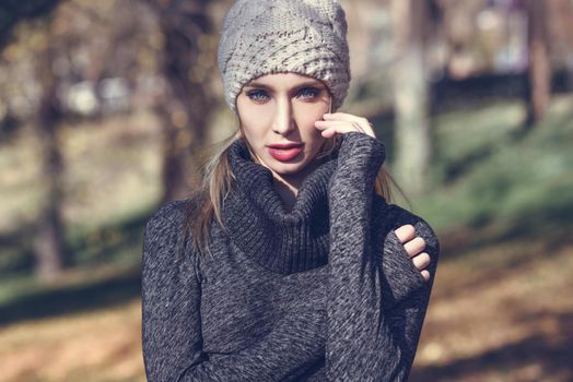 Close-up portrait of young blonde woman standing in a park with autumn colors. Beautiful girl wearing winter gray dress and wool cap. Female with straight hair and blue eyes.