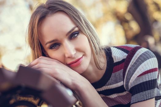 Close-up portrait of young blonde woman sitting on a bench in the street of a park with autumn colors. Beautiful girl with blue eyes in urban background wearing striped dress smiling.