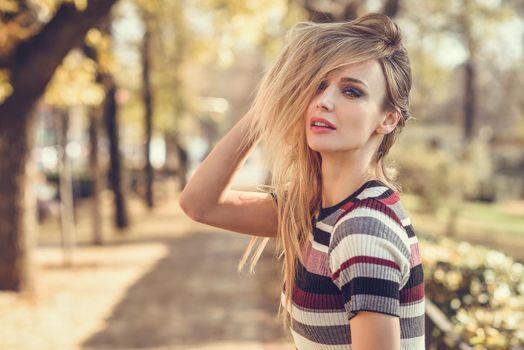 Young blonde woman standing in the street moving her hair. Beautiful girl in urban background wearing striped dress. Female with straight hair and blue eyes.
