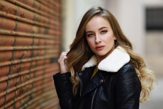 Blonde woman in urban background. Beautiful young girl wearing black leather jacket and mini skirt standing in the street. Pretty russian female with long wavy hair hairstyle and blue eyes.