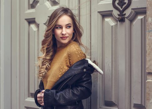 Blonde woman in urban background. Beautiful young girl wearing black leather jacket and mini skirt standing in the street. Pretty russian female with long wavy hair hairstyle and blue eyes.
