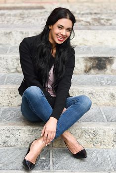 Portrait of hispanic young woman smiling and wearing casual clothes sitting on steps in urban background