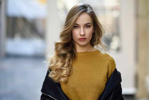 Blonde woman in urban background. Beautiful young girl wearing black leather jacket and mini skirt standing in the street. Pretty russian female with long wavy hair hairstyle and blue eyes.