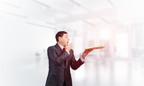 Scared businessman covering mouth with hand and holding open book. Startled adult man in business suit and tie standing on blurred office background. Education and knowledges. Business accounting