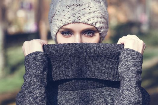 Close-up portrait of young blonde woman standing in a park with autumn colors. Beautiful girl wearing winter gray dress and wool cap. Female with straight hair and blue eyes.