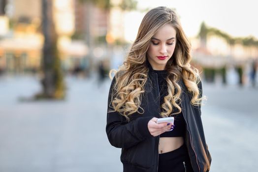 Blonde woman texting with her smart phone in urban background. Beautiful young girl wearing black jacket walking in the street. Pretty russian female with long wavy hair hairstyle.