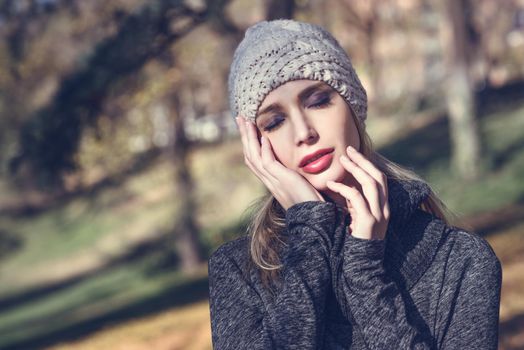 Young blonde woman standing in a park with autumn colors. Beautiful girl wearing winter gray dress and wool cap. Female with straight hair and eyes closed with dark eyeshadow.