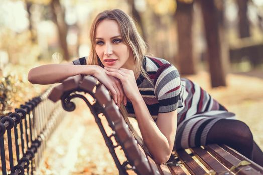 Pensive young blonde woman sitting on a bench in the street of a park with autumn colors. Beautiful girl in urban background wearing striped dress and black tights. Female with straight hair and blue eyes.