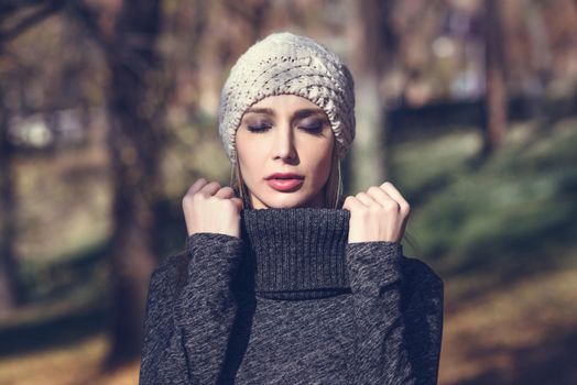 Young blonde woman standing in a park with autumn colors. Beautiful girl wearing winter gray dress and wool cap. Female with straight hair and eyes closed with dark eyeshadow.