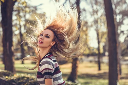 Young blonde woman standing in the street moving her hair. Beautiful girl in urban background wearing striped dress. Female with straight hair and blue eyes.