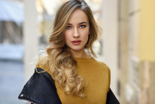 Blonde woman in urban background. Beautiful young girl wearing black leather jacket and mini skirt standing in the street. Pretty russian female with long wavy hair hairstyle and blue eyes.