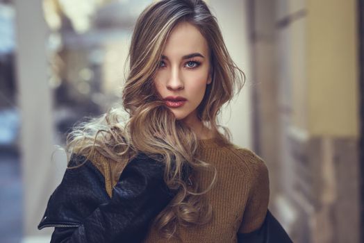 Blonde woman in urban background. Beautiful young girl wearing black leather jacket and mini skirt standing in the street. Pretty russian female with long wavy hair hairstyle and blue eyes.