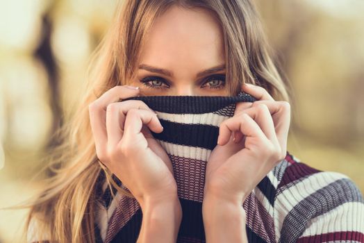 Close-up portrait of young blonde woman with blue eyes standing in the street. Beautiful girl in urban background wearing striped sweater. Female with straight hair.