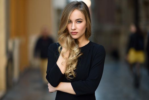 Blonde woman in urban background. Beautiful young girl wearing black elegant dress standing in the street. Pretty russian female with long wavy hair hairstyle and blue eyes.