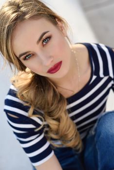 Close-up portrait of blonde woman, model of fashion, possing in urban background. Beautiful young girl wearing striped t-shirt and blue jeans in the street. Pretty russian female with pigtail.
