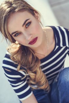 Close-up portrait of blonde woman, model of fashion, possing in urban background. Beautiful young girl wearing striped t-shirt and blue jeans in the street. Pretty russian female with pigtail.