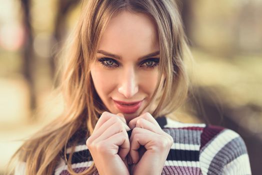 Close-up portrait of young blonde woman with blue eyes standing in the street. Beautiful girl in urban background wearing striped sweater. Female with straight hair.