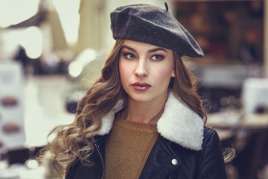 Blonde russian woman in urban background. Beautiful young girl wearing beret, black leather jacket and mini skirt standing in the street. Pretty female with long wavy hair hairstyle and blue eyes.