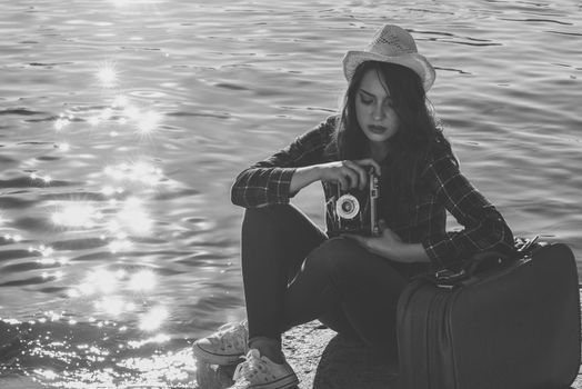Young woman taking photographs with a vintage camera sitting in a harbour. Girl wearing plaid shirt, blue jeans and sun hat. Black and white photograph.
