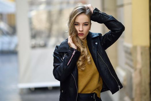 Blonde woman in urban background. Beautiful young girl wearing black leather jacket and mini skirt standing in the street. Pretty russian female with long wavy hair hairstyle and blue eyes.