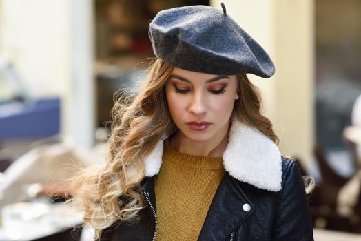 Blonde russian woman in urban background. Beautiful young girl wearing beret, black leather jacket and mini skirt standing in the street. Pretty female with long wavy hair hairstyle.