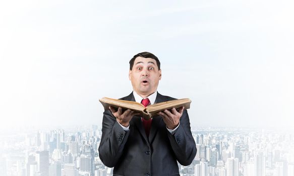 Surprised businessman holding open old book. Startled adult man in business suit and tie standing on downtown background. Business education and knowledges. Business paperwork and bookkeeping concept.
