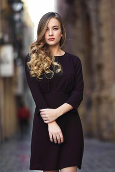 Blonde woman in urban background. Beautiful young girl wearing black elegant dress standing in the street. Pretty russian female with long wavy hair hairstyle and blue eyes.