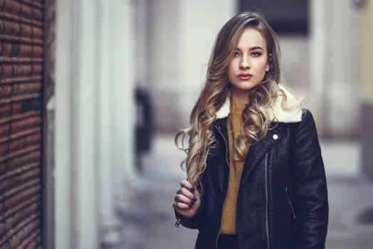 Blonde woman in urban background. Beautiful young girl wearing black leather jacket and mini skirt standing in the street. Pretty russian female with long wavy hair hairstyle and blue eyes.