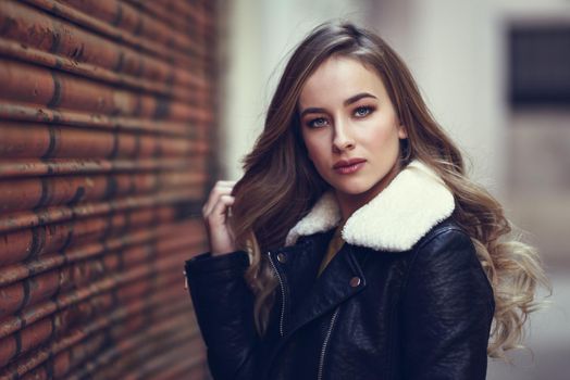 Blonde woman in urban background. Beautiful young girl wearing black leather jacket and mini skirt standing in the street. Pretty russian female with long wavy hair hairstyle and blue eyes.